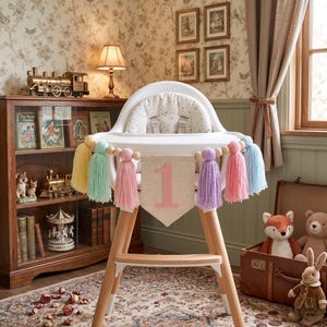 May include: A white high chair with wooden legs and a banner that says "1". The banner is decorated with colorful tassels. The high chair is set in a room with a bookshelf, toys, and a window.