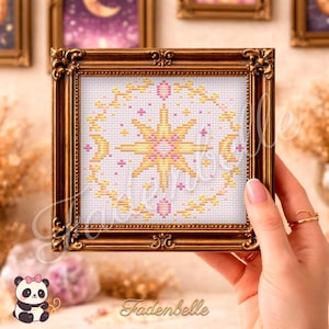May include: A framed cross-stitch artwork with a celestial design. The design includes a central star, crescent moons, and floral elements in yellow and pink. The ornate, gold-coloured frame surrounds the artwork, which is signed "Fadenbelle".
