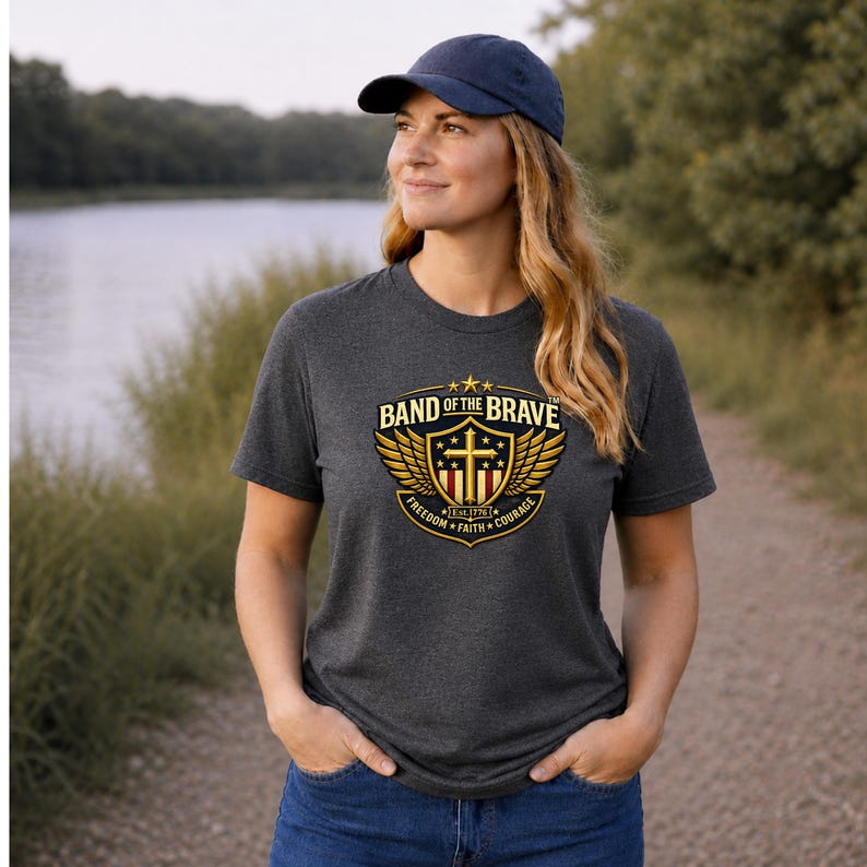 May include: A dark charcoal gray short-sleeve t-shirt featuring a gold and red graphic emblem with wings, a cross, and the text "BAND OF THE BRAVE" above "FREEDOM FAITH COURAGE" and "EST. 1776". A navy blue baseball cap is worn with the t-shirt.