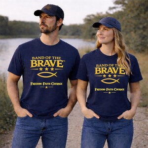 May include: A navy blue t-shirt featuring the yellow text BAND OF THE BRAVE, three stars, a fish symbol, and the words FREEDOM FAITH COURAGE with a star below. The shirt is paired with a matching navy blue baseball cap worn on the head.