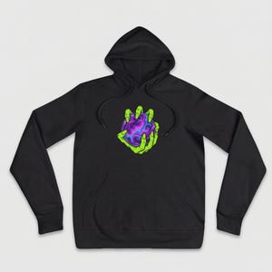 Puede incluir: Sudadera con capucha negra con bolsillo delantero y cordón. El gráfico en la parte delantera presenta manos verdes con garras que sostienen un orbe morado y azul. El diseño tiene un tema de fantasía.