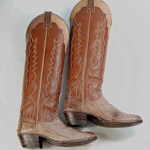 Puede incluir: Un par de botas de vaquero de cuero marrón con costuras decorativas y un patrón de piel de serpiente en la parte inferior. Las botas tienen una punta puntiaguda y un tacón apilado. Las cañas de las botas son altas.