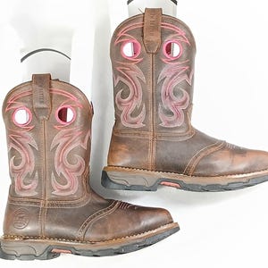 May include: A pair of brown leather cowboy boots with pink accents. The boots feature decorative stitching in pink and a sturdy sole. The top of the boots have the text "IRISH SETTER".