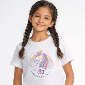 Peut inclure: Un t-shirt blanc à manches courtes orné d'une licorne à la crinière arc-en-ciel, portant une couronne dorée au-dessus de nuages. Le texte Little Kaur, Big Dreams est inscrit avec le mot Kaur en écriture gurmukhi violette sous la licorne.