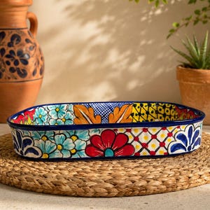 May include: An oval, hand-painted ceramic baking dish with a vibrant floral design. The dish features a blue rim and a colorful pattern of flowers and geometric shapes in red, yellow, and turquoise. It sits on a woven mat.