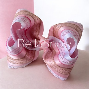 May include: A large, layered hair bow in shades of pink, white, and gold. The bow features a mix of satin and lace ribbons, with the text "BellaSnow" across the front. The bow is designed with multiple loops and a central knot.