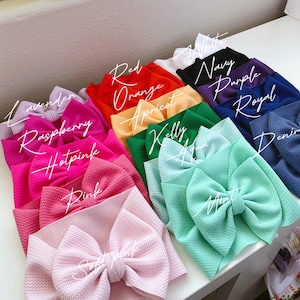 May include: A collection of colorful baby headbands in various shades, including pink, red, orange, green, and blue. Each headband features a large bow and is labeled with its color name in cursive script. The headbands are arranged on a white surface.