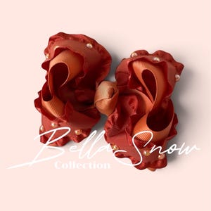 May include: A rust-colored hair bow featuring ruffled edges and small embedded pearls. The center is gathered with a matching ribbon. The text Bella Snow Collection is overlaid in white script across the center of the bow.