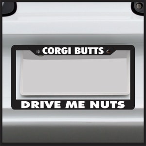 May include: Black license plate frame with the words "CORGI BUTTS" above and "DRIVE ME NUTS" below in white capital letters. The frame surrounds a blank white license plate.