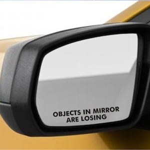 May include: A black car side mirror with a white sticker that reads "Objects in mirror are losing".
