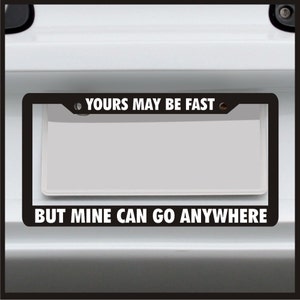 May include: Black license plate frame with white text that reads "YOURS MAY BE FAST" at the top and "BUT MINE CAN GO ANYWHERE" at the bottom. The frame is mounted on a white vehicle.