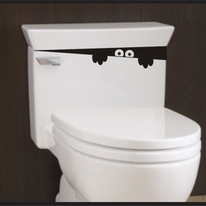 May include: A white toilet with a black and white sticker of a monster peeking over the top of the toilet tank. The monster has two eyes and two hands.