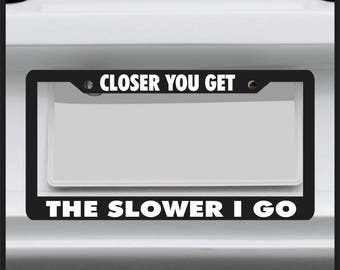 May include: Black license plate frame with white text that reads "CLOSER YOU GET THE SLOWER I GO".