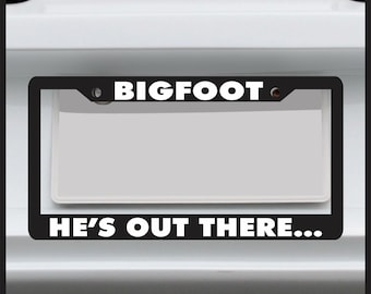 Bigfoot He’s Out There Funny License ‑ Plate Frame — Fits US/Canada Tags, ABS Plastic, Black w/ White Text, Gift for Sasquatch Fans