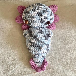 May include: A handmade crochet axolotl plushie in shades of blue, grey, and white, with pink gills and tail. The axolotl has brown wooden bead eyes. The plushie is lying on a white surface.
