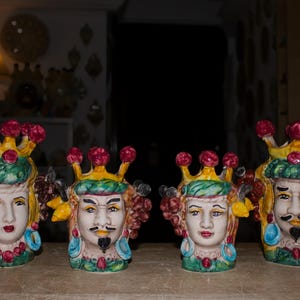 Könnte beinhalten: Vier Keramikkopfskulpturen mit Kronen, die detaillierte Gesichter und farbenfrohe Akzente aufweisen. Die Skulpturen haben gelbe, grüne, rote und blaue Details. Die Köpfe sind in einer Reihe auf einer Holzoberfläche angeordnet.