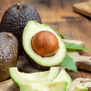May include: Close-up of fresh avocados, one halved to reveal the large seed. The green flesh contrasts with the dark, textured skin. Sliced avocado pieces are also visible, suggesting a culinary theme.
