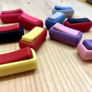 May include: A collection of colorful, rectangular 3D-printed objects with a sliding top. The objects are in various colors, including red, pink, yellow, blue, and purple. The items are arranged on a light-colored wooden surface.
