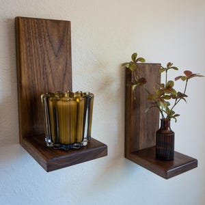 May include: Two wooden floating shelves with a dark finish are mounted on a white wall. One shelf holds a glass candle holder with a lit candle, and the other holds a small brown vase with greenery. The shelves are from Finney Woodworks.