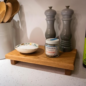 May include: A wooden kitchen countertop display with a small ceramic bowl filled with salt, a jar of Jacobsen Co. Loveski seasoning, two gray pepper grinders, and a bottle of olive oil. Wooden spoons are in a white container.