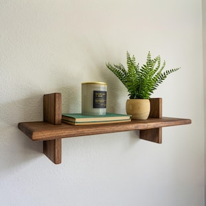 May include: A dark wood floating shelf with a candle, a book, and a potted fern. The shelf is mounted on a white wall. The candle has a gold lid and a black label. The book is green. The fern is in a small, light-colored pot.