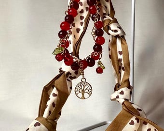 May include: A scarf with a brown and white heart pattern is adorned with a red beaded bracelet. The bracelet features a gold tree charm and small green leaf accents. The scarf is knotted and hangs from a gold ring.