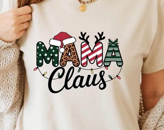 May include: A cream-colored t-shirt with the words "Mama Claus" in a festive design. The letters are decorated with polka dots, leopard print, stripes, and a bow. A Santa hat and reindeer antlers are incorporated into the design, with a string of lights below.