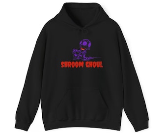 May include: A black hooded sweatshirt featuring a graphic of a purple mushroom-headed creature with a wide orange grin. Below the creature, the text SHROOM GHOUL is printed in bold, bright orange capital letters. The hoodie has long sleeves.
