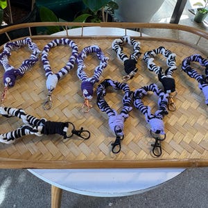 May include: A collection of handmade lanyards with a braided design. The lanyards feature a black and white pattern, with accents of purple and black. Each lanyard has a metal clasp for attaching keys or a badge. They are displayed on a woven tray.