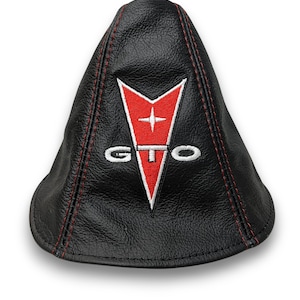 May include: Black shift boot with red stitching. Features a red and white Pontiac GTO emblem. The emblem is a red arrowhead shape with a white star and the letters "GTO" in white.