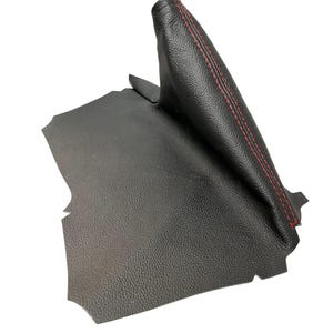 Könnte beinhalten: Schwarzer Leder-Handbremsbezug für Autos mit roten Nähten. Der Bezug hat eine strukturierte Oberfläche und ist für den Handbremshebel konzipiert. Ein passendes Lederstück ist ebenfalls zu sehen.