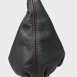 Op de afbeelding: Zwarte leren versnellingspookhoes met rode stiksels. De hoes heeft een textuur en een conische vorm. De rode stiksels lopen langs de naad van de hoes en zorgen voor een contrasterend detail.