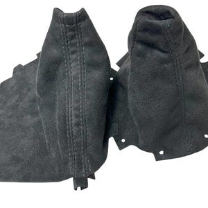 Könnte beinhalten: Zwei schwarze Wildleder-Schaltmanschetten, eine mit einer genähten Naht in der Mitte und die andere mit einem abgerundeten Oberteil. Die Manschetten haben eine strukturierte Oberfläche und sind für die Montage um den Schalthebel eines Fahrzeugs konzipiert.