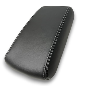 Puede incluir: Apoyabrazos automotriz negro y rectangular con costuras blancas. El apoyabrazos tiene una superficie ligeramente texturizada y bordes redondeados. Las costuras recorren el perímetro, creando un borde definido.