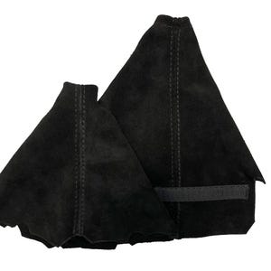 Peut inclure: Deux couvre-bottes de changement de vitesse en daim noir. Les housses sont de forme triangulaire, avec un bord festonné sur l'une et un bord droit sur l'autre. Chaque housse a une couture verticale avec des coutures visibles. Une bande velcro noire est attachée.