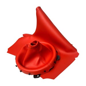 Puede incluir: Un fuelle de palanca de cambios de coche y un panel de cuero rojo brillante. El fuelle es circular en la base y se estrecha hacia arriba. El panel de cuero es rectangular con un borde curvo. Un accesorio interior de coche.