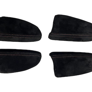May include: Four black automotive armrest covers featuring a soft, suede-like texture and prominent red stitching along the edges. These contoured covers are designed to fit vehicle interior components, showcasing a uniform dark finish and detailed seams.