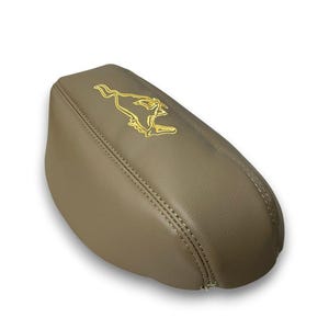 Puede incluir: Una funda acolchada para reposabrazos de coche de color beige con un logotipo de caballo corriendo bordado en dorado. La funda tiene un detalle de costura y está diseñada para adaptarse a la consola central de un coche. La funda es de material liso similar al cuero.