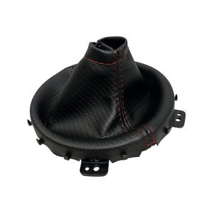 May include: A black gear shift boot with red stitching. The boot has a textured, carbon fibre-like appearance and a circular base with mounting tabs. The stitching is visible along the seams, adding a contrasting detail to the design.