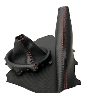 Op de afbeelding: Zwarte leren versnellingspookhoes en versnellingspookknophoes met rode stiksels. De versnellingspookhoes heeft een kegelvorm en de versnellingspookknophoes een afgeronde vorm. De items liggen op een zwart oppervlak met textuur.