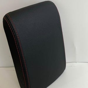 May include: A black, rectangular cushion with rounded edges. The cushion has a textured surface and is stitched with red thread along the seams. The item appears to be made of leather or a leather-like material.