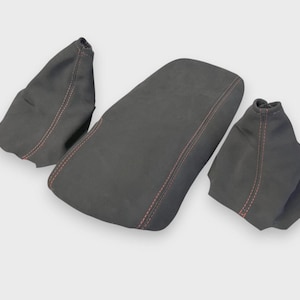 May include: A set of three dark gray automotive interior pieces. The pieces include a center console cover and two shift boot covers. Each piece has red stitching for a contrasting accent.