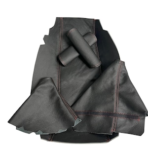 Op de afbeelding: Zwarte lederen auto-interieuraccessoires met rode stiksels. De set bevat een versnellingspookhoes, twee armleuninghoezen en andere onderdelen. Het leer heeft een getextureerde uitstraling.