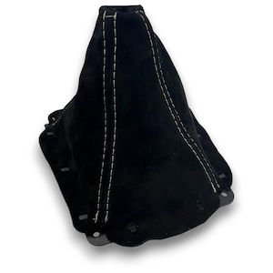 Puede incluir: Fuelle de palanca de cambios de ante negro con costuras blancas. La base acampanada tiene orificios de montaje. Las costuras blancas contrastan con el material oscuro, resaltando las costuras.