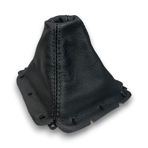 May include: A black leather shift boot with a textured surface. The boot has a stitched seam down the center and small tabs around the base. The product is designed to fit around a vehicle's gear shift lever.
