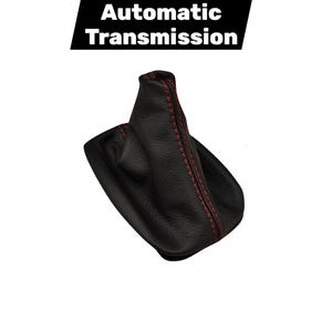 Puede incluir: Fuelle de palanca de cambios automática de cuero negro con costuras rojas. El texto "Automatic Transmission" se muestra en un rectángulo negro encima del fuelle. El fuelle está diseñado para cubrir la palanca de cambios.