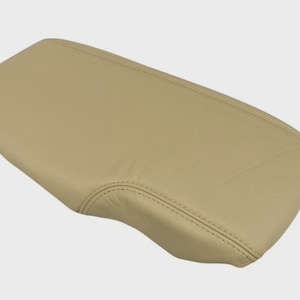 May include: A tan, rectangular car armrest cover. The cover is made of a smooth material and features stitched detailing along the edges. The armrest cover is designed to fit a car's center console.