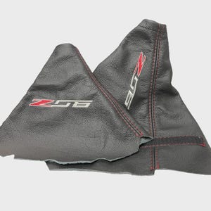 May include: Two black leather shift boots with red stitching. One boot has "Z06" in red and silver lettering, and the other has "Z06" in red and silver lettering. The boots are designed for a vehicle's gear shift.
