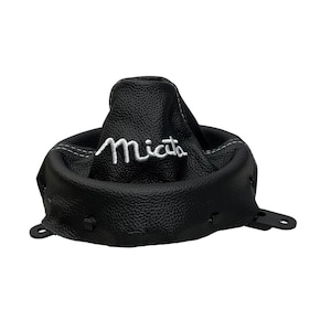 May include: A black leather gear shift boot with a textured surface and white stitching. The word Miata is embroidered in white cursive script on the front panel. The boot features a circular base with integrated black plastic mounting tabs.