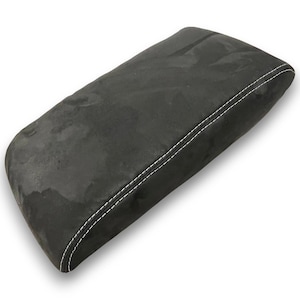 Puede incluir: Un reposabrazos de coche negro y rectangular con costuras blancas. El reposabrazos tiene un aspecto texturizado, similar al ante. Está diseñado para el interior de un vehículo.